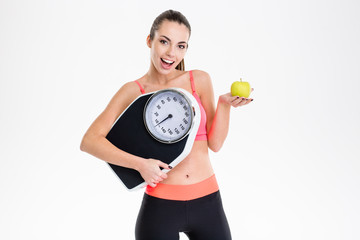 Wall Mural - Excited positive fitness girl holding weighing scale and apple