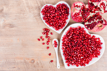 Two white heart shaped plates full of fresh ripe juicy pomegranate seeds, little spoon, whole fruit and ripe one on wooden background.