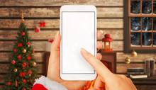Woman With Smart Phone In Hands In Christmas Time. Device With White Screen For Mockup. Christmas Tree, Gifts, Decorations In Background.