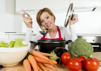 young happy home cook woman in red apron at domestic kitchen holding saucepan tasting hot soup