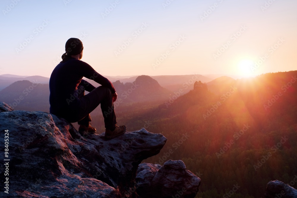 Tired adult hiker in black trousers, jacket and dark cap sit on cliff edge and looking to colorful mist in valley bellow - obrazy, fototapety, plakaty 