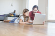 © Westend61 - Mother and daughter lying on floor using laptop, father making music in background