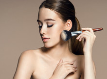Young woman applying blusher on her face with powder brush, skin care concept / photo composition of brunette girl - on beige background