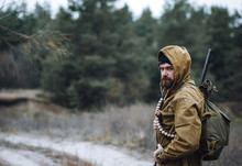 Brutal Hunter, Bearded Man In Warm Hat With A Gun In His Hand, A Knife A Backpack And Smoking Pipe In The Wild Forest In The Autumn