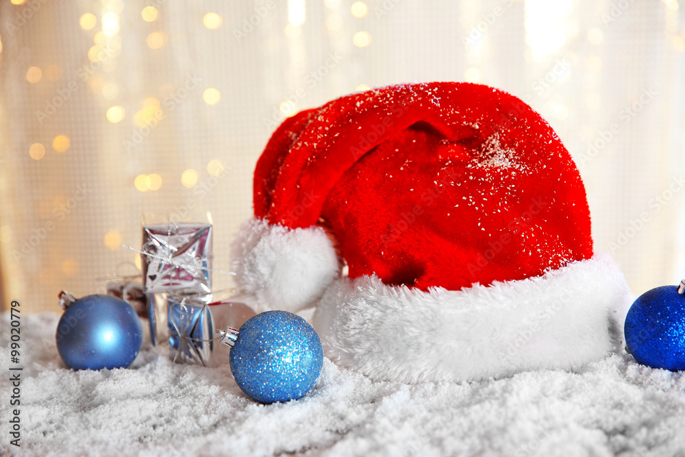Santa Claus hat with baubles and gift boxes on a snowy table over glitter background