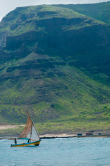 Small wooden fisherman sail boat with two men in the blue windy ocean front of a green mountain