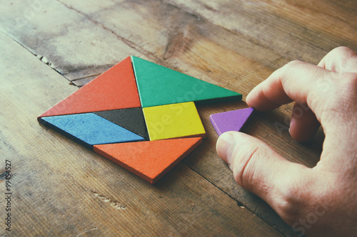 Fotografie, Obraz man's hand holding a missing piece in a square tangram puzzle, over wooden table