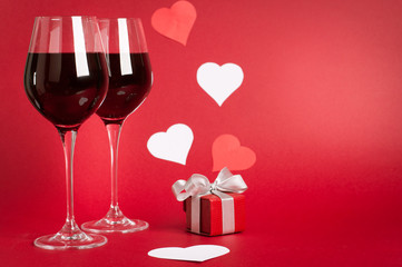 Valentine’s day hearts, gift box and wine glasses. Red matte background. White and red elements. 