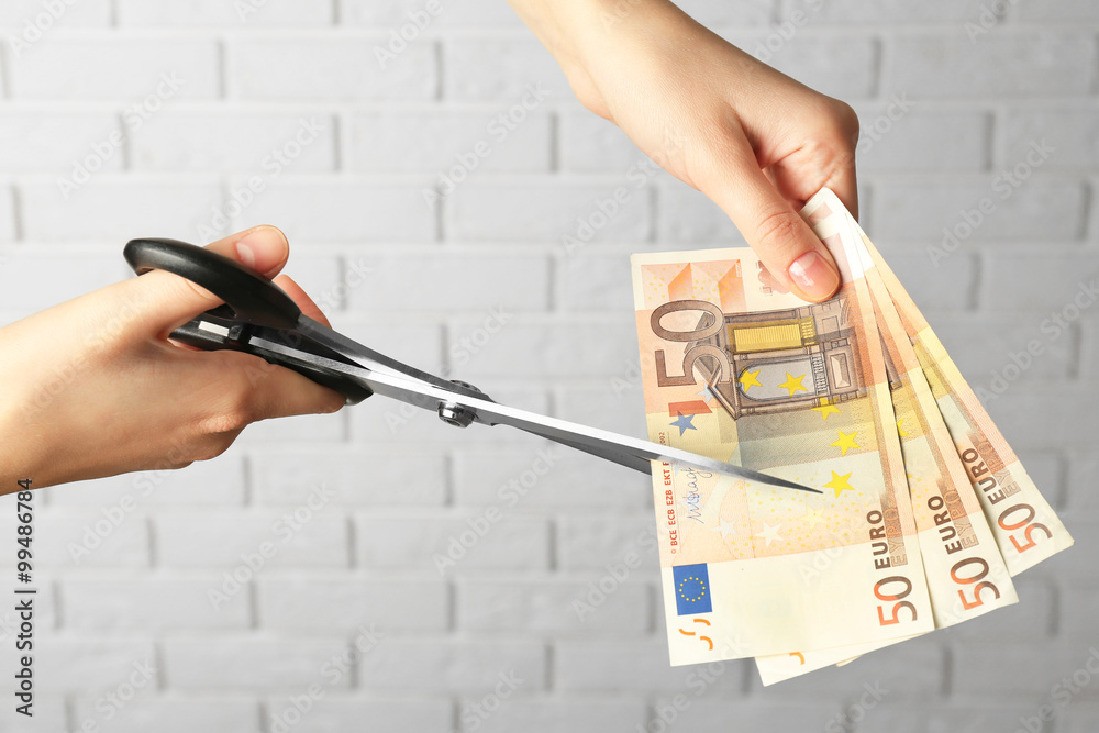 Hands with scissors cutting money on white brick wall background