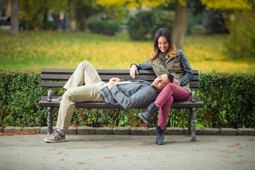 Wall Mural - Happy young woman sitting on a bench in a park with her boyfriend lying on her lap