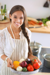 Wall Mural - Smiling young woman holding vegetables standing in kitchen