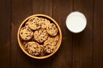 Wall Mural - Chocolate chip cookies in wooden bowl with a glass of cold milk on the side photographed on wood with natural light