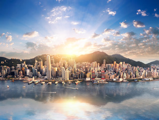 Hong Kong city skyline view from harbor with skyscrapers buildings reflect in water at sunset with sunlight and sun rays shine through clouds on blue sky