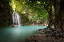 Sunlight Beams And Rays Shine Through Leaves Of Trees In Tropical Rainforest Park In Thailand With Beautiful Waterfall Falling In Clear Pond And Old Big Tree On Foreground