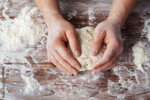 Fotografie, Obraz Baker prepares the dough on a wooden table, male hands knead the dough with flou