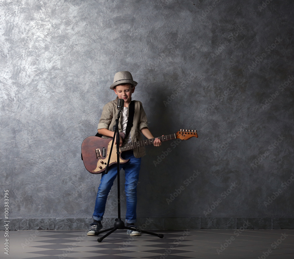 Little boy playing guitar and singing with microphone on a grey wall background