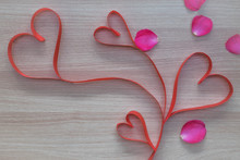 Four Red Heart Shape Ribbon With Pink Rose Petals On Wooden Surface With Empty Space For Text