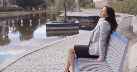 Wall Mural - Business woman with peaceful expression sitting back to relax outdoors on bench near canal