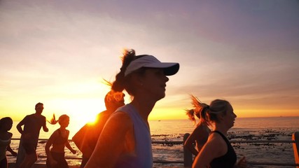 Wall Mural - Group of runners running on promenade by the sea in sunset. Healthy young people training together outdoors in evening.
