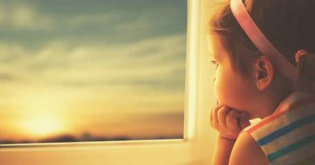 Wall Mural - child sad little girl looking out window at sunset