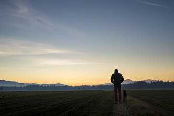 View from behind of a man walking with his black dog at dusk on