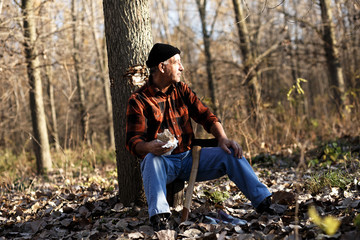 Poster - Portrait of senior lumberjack in forest sitting on a tree stump. He is resting and having lunch break.