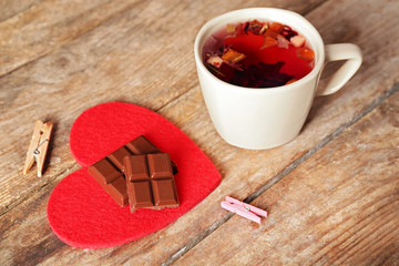 Cup of fruit tea with piece of chocolate on red heart and clothespins on wooden background closeup