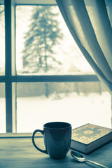 Cup of coffee and book near window on a snowy winter day