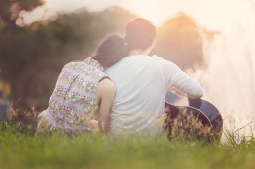 Lovers in a lake. Young couple in love sitting on the park ground near the water while these young man playing guitar in sunset time. Post processing in vintage style