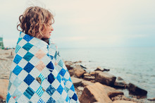 Cute Curly Happy Child Girl Relaxing On Stone Beach, Wrapped In Cozy Quilt Blanket. Summer Vacation Activities, Traveling In Europe On Holidays.