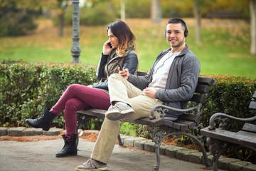 Wall Mural - Young man listening to music while woman sitting on the same park bench is talking on the phone