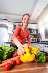 Wall Mural - Wide angle view of young woman making healthy food standing happy smiling in kitchen holding pepper. Focus on pepper.