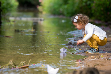 Wall Mural - Cute little girl runs a paper boat in the stream in the park. Stretching her hand and reaching the little ship