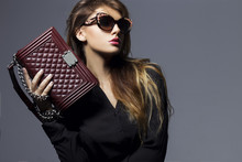 Portrait Of A Beautiful Girl Posing In Studio In Black Shirt, Blue Jeans And Fashion Sunglasses, Holding Burgundy Handbag . The Concept Of Stylish And Sexy Women. Perfect Skin And Body