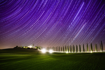 Beautiful Tuscany night landscape with star trails on the sky, cypresses and shining road in green meadow. Natural outdoor amazing fantasy background.