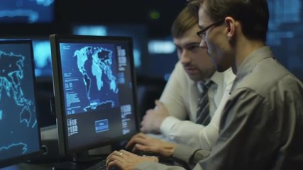 Two IT programmers are working on computer in a dark office room filled with display screens. Shot on RED Cinema Camera.