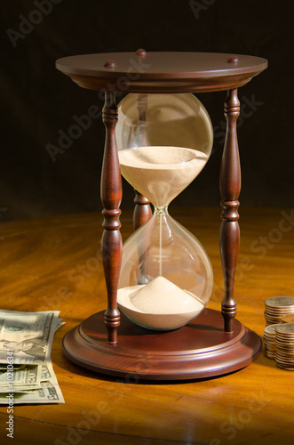 Fényképezés  Large hour glass surrounded by paper cash money and coins on a wooden table representing time running out,  investing time, deadlines, stress, financial planning and the wisdom that comes with time