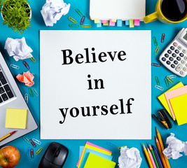 Believe in yourself. Office table desk with supplies, white blank note pad, cup, pen, pc, crumpled paper, flower on wooden background. Top view