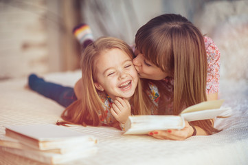 Portrait of a smiling young cute mother and daughter reading a book lying and relax in the bed in a bright big white room 