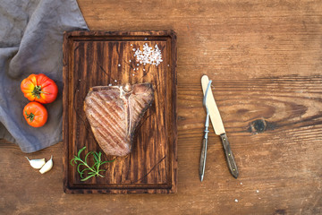 Wall Mural - Cooked meat t-bone steak on serving board with garlic cloves, tomatoes, rosemary and spices over rustic wooden background.