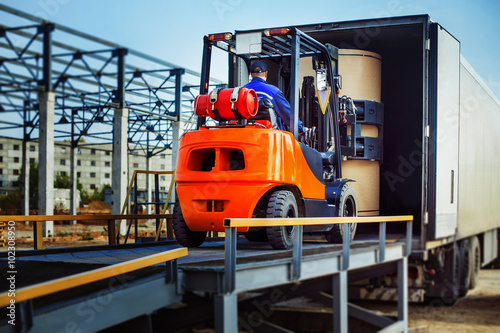 Fotografie, Obraz Forklift is putting cargo from warehouse to truck outdoors