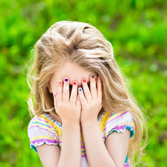 Wall Mural - Beautiful little girl with long blond hair and many-coloured manicure covering her eyes with her hands