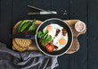 Pan of fried eggs, bacon, tomatoes with bread, mangold and cucumbers on rustic wooden serving board over dark table surface