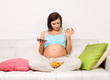 Pregnant happy smiling woman sitting on a sofa eating unhealthy junk food snack. Young beautiful mom expecting Baby. Pregnant Woman Belly. Maternity concept.