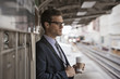 © Mint Images - A working day. Businessman in a work suit and tie holding a cup of coffee on a railway platform.