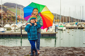 Wall Mural - Outdoor portrait of two cute kids resting by the lake on a rainy day, hiding under big colorful umbrella