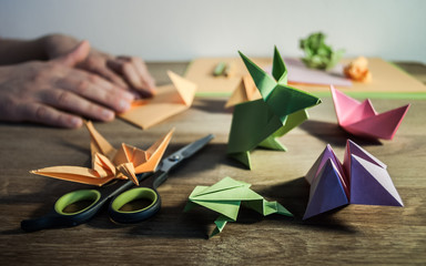 origami figures, scissors and pencils on wooden table, in the background hands folding colored paper