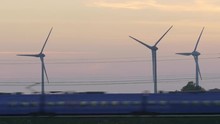 A Passenger Train Goes By At Dusk In A Rural Countryside Setting With Energy Producing Wind Turbines And Power Lines In The Background. Closeup. Location: Lund, Sweden (Scania) In June 2015.
