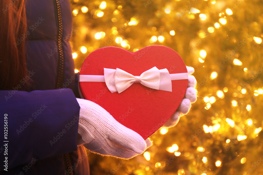 Red heart shaped box with bow in hands over lighted background