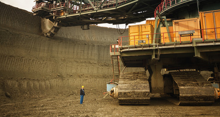 Coal mine worker with a helmet on his head standing in front of huge drill machine and looking at it.